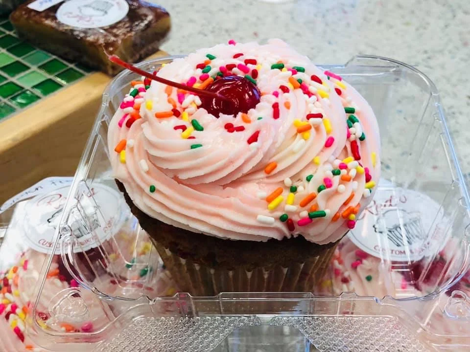 A pink-frosted cupcake with rainbow sprinkles and a cherry on top